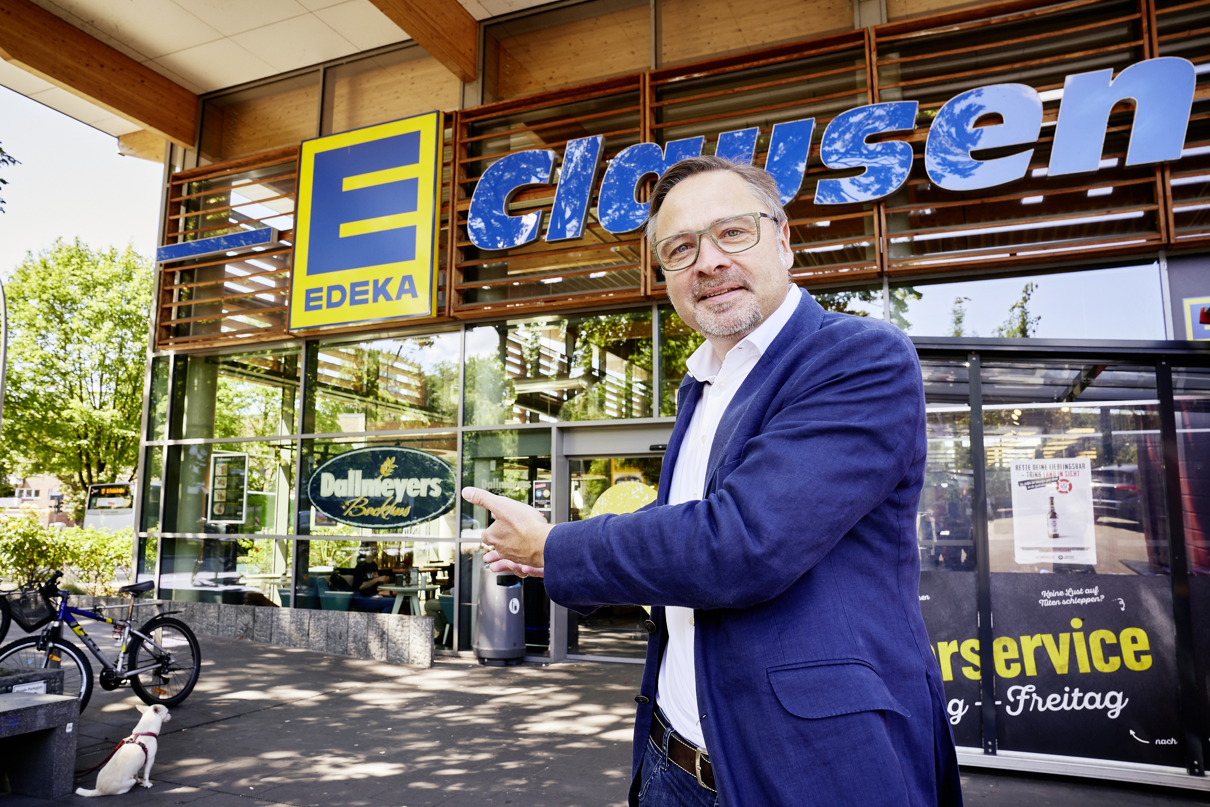 Ein Mann mit Brille und in blauem Sakko steht vor EDEKA Clausen. Er hat beide Handflächen nach oben gerichtet.