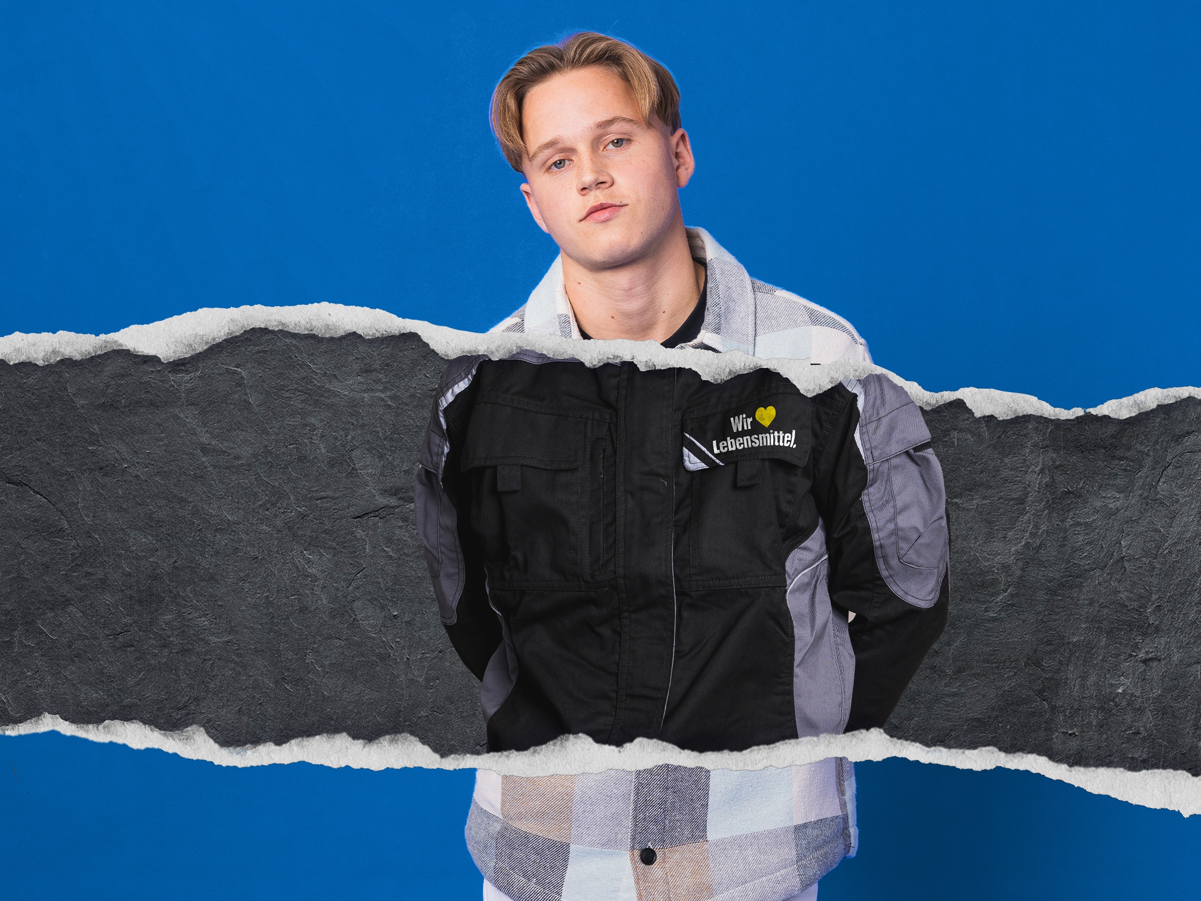 Junger blonder Mann mit Mittelscheitel trägt einen schwarzen Arbeitsanzug mit der Aufschrift Wir lieben Lebensmittel.