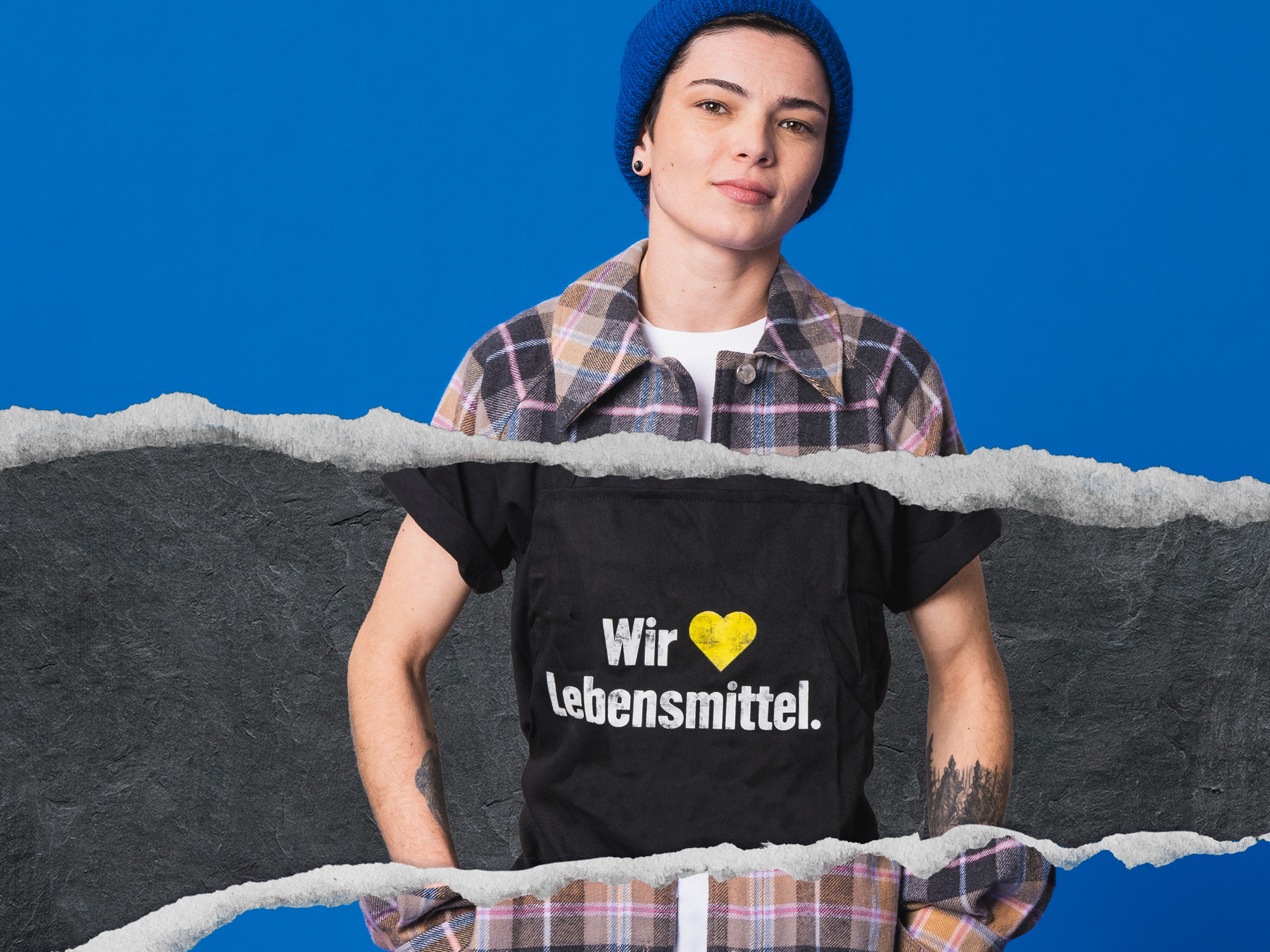    	 Junge Person mit blauer Wollmütze, Karohemd und tätowierten Unterarmen trägt eine schwarze Schürze.