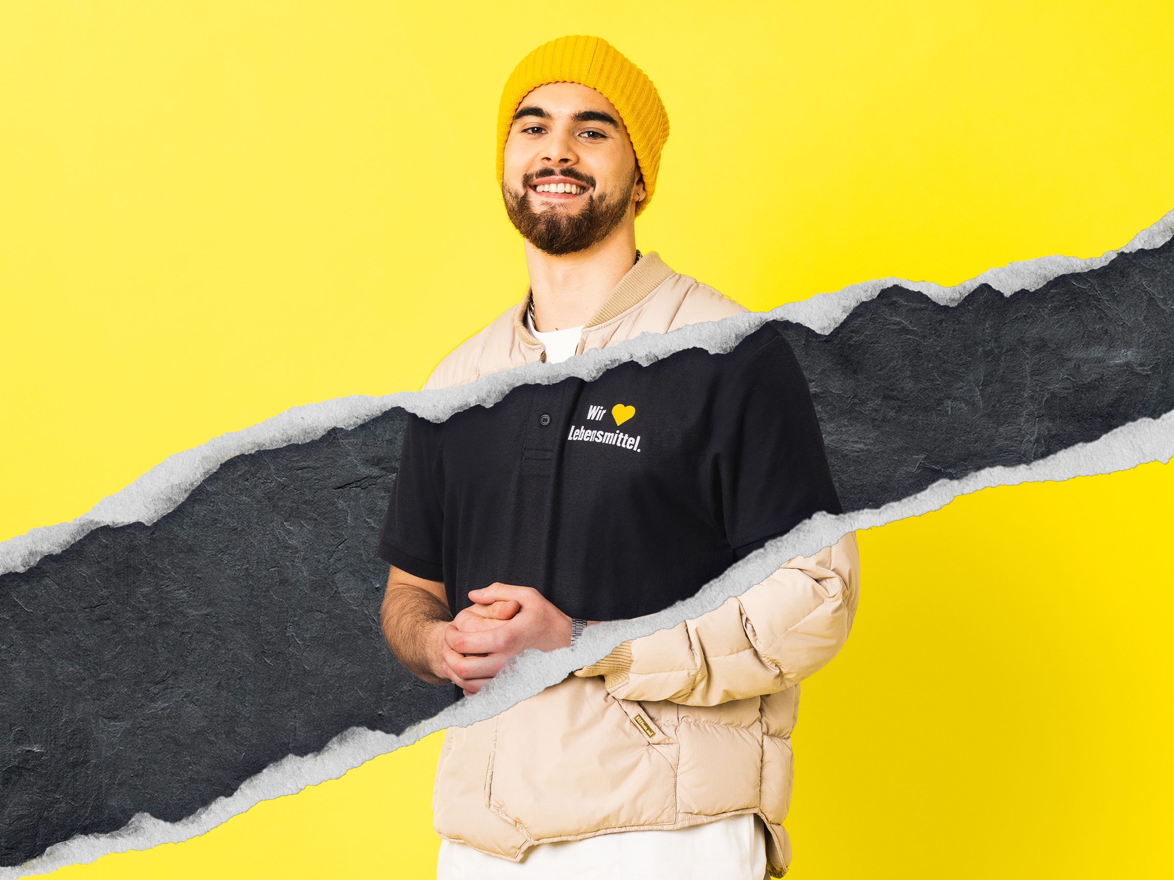 Junger Mann mit dunklem Vollbart und gelber Wollmütze trägt ein Poloshirt mit der Aufschrift Wir lieben Lebensmittel.