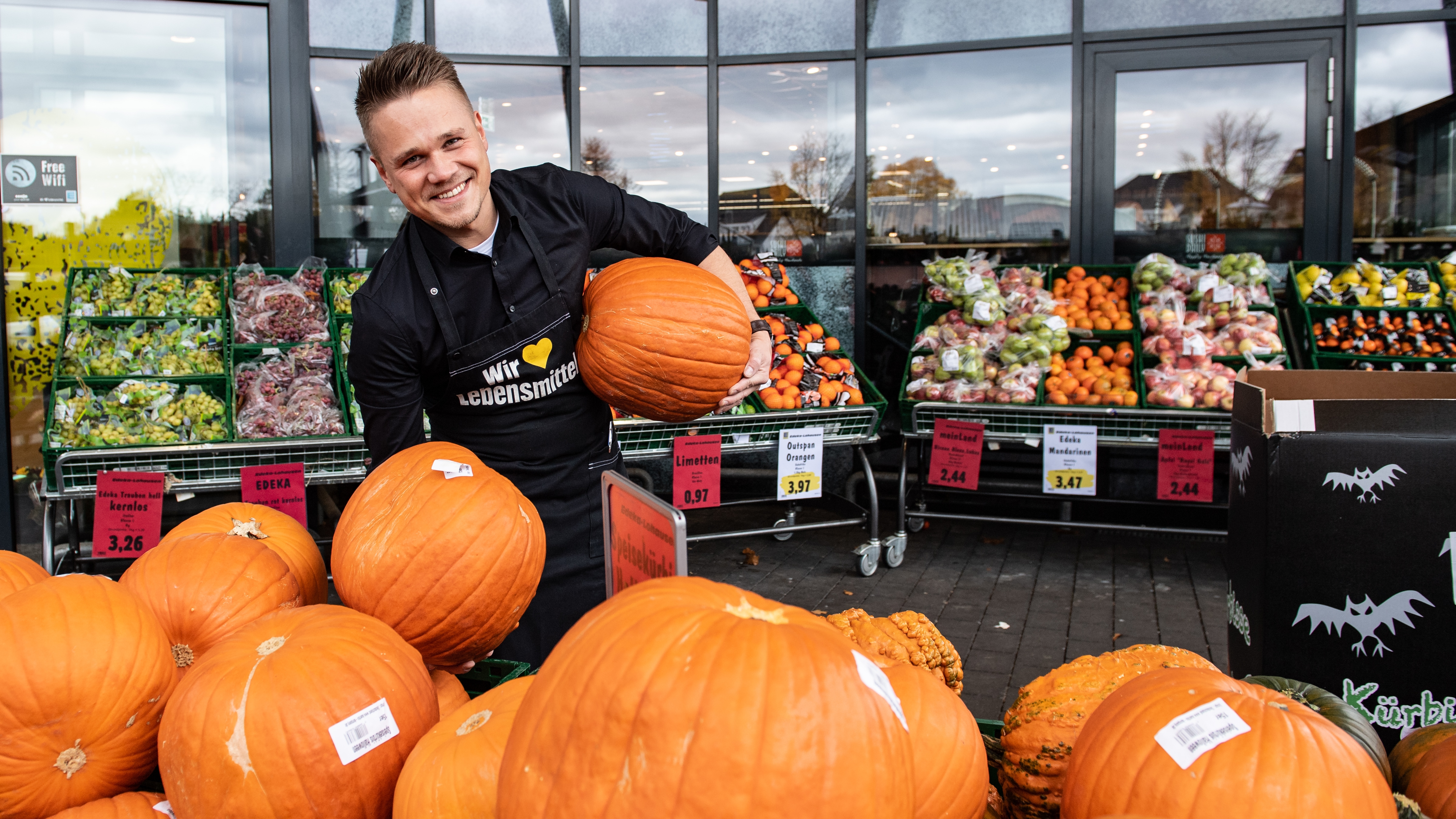 Marvin Waschkuhn vor einem EDEKA-Markt. Er hält in jeder Hand einen Riesenkürbis, trägt eine schwarze EDEKA-Schürze und lacht
