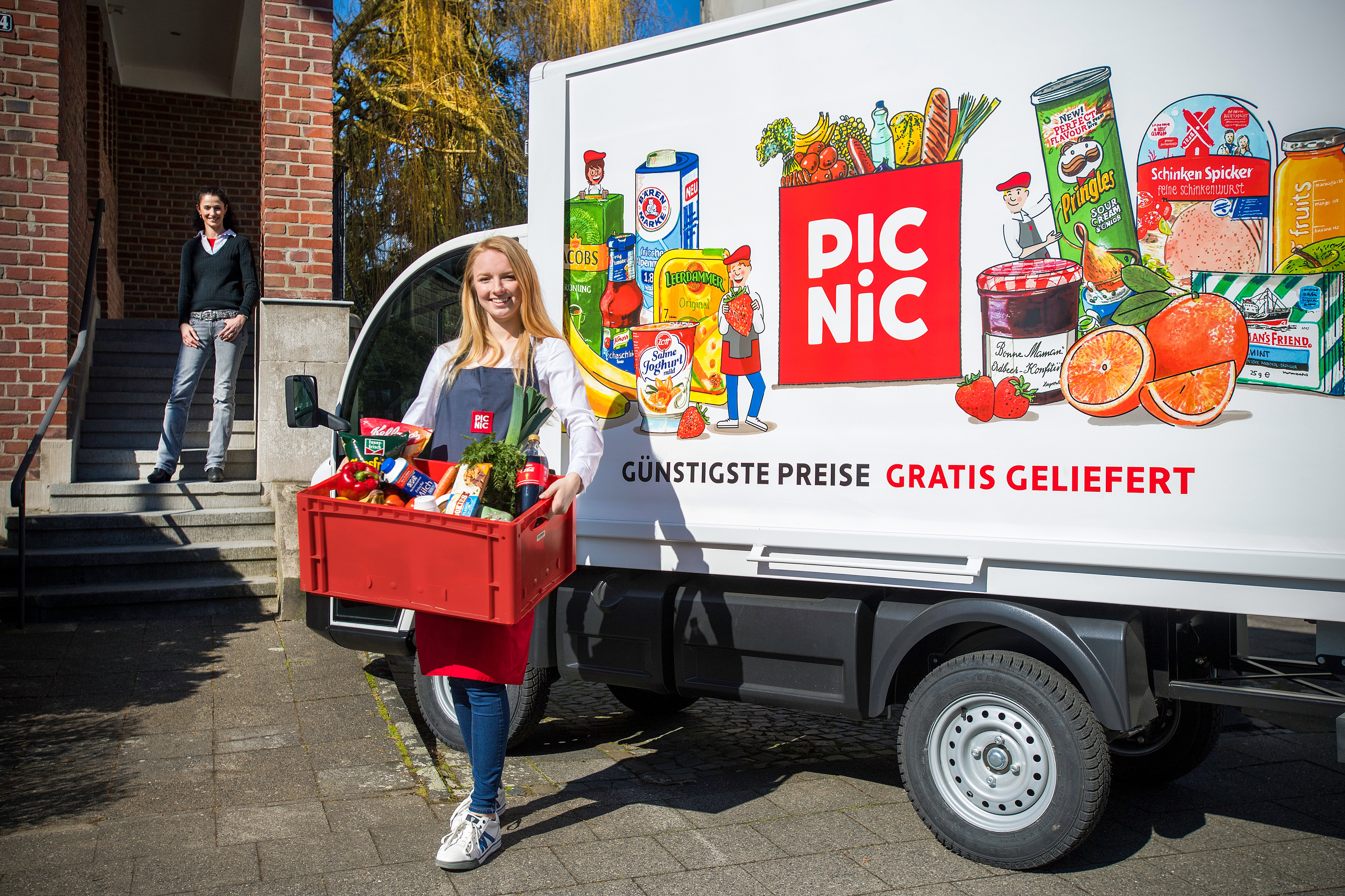    	 Eine blonde Frau mit einer Schürze trägt eine rote Kiste mit Lebensmitteln. Sie steht vor einem Liefervan vpn Picnic.