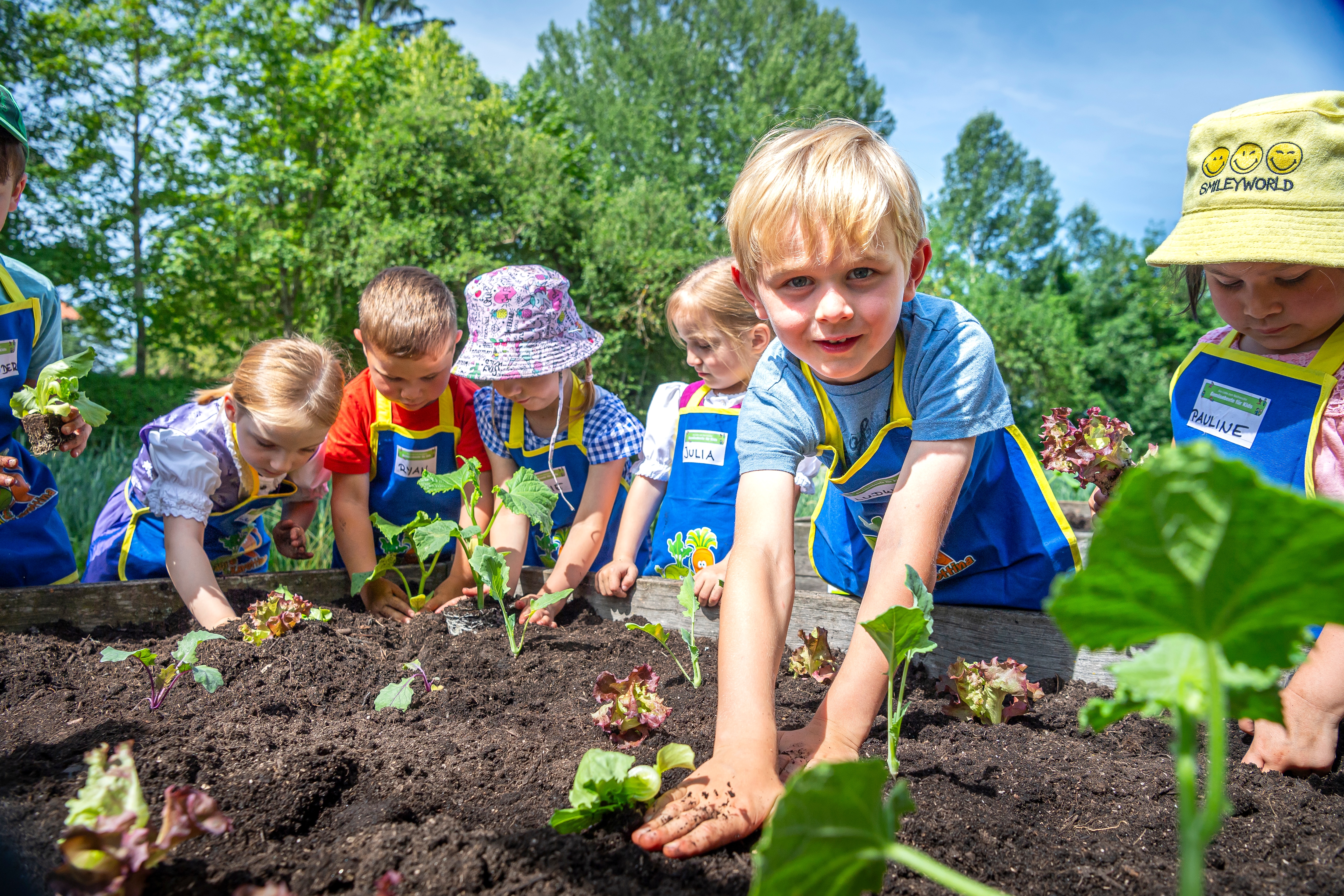 Sechs kleine Kinder stehen an einem Hochbeet. Sie tragen Schürzen und stecken die Hände in die Erde.