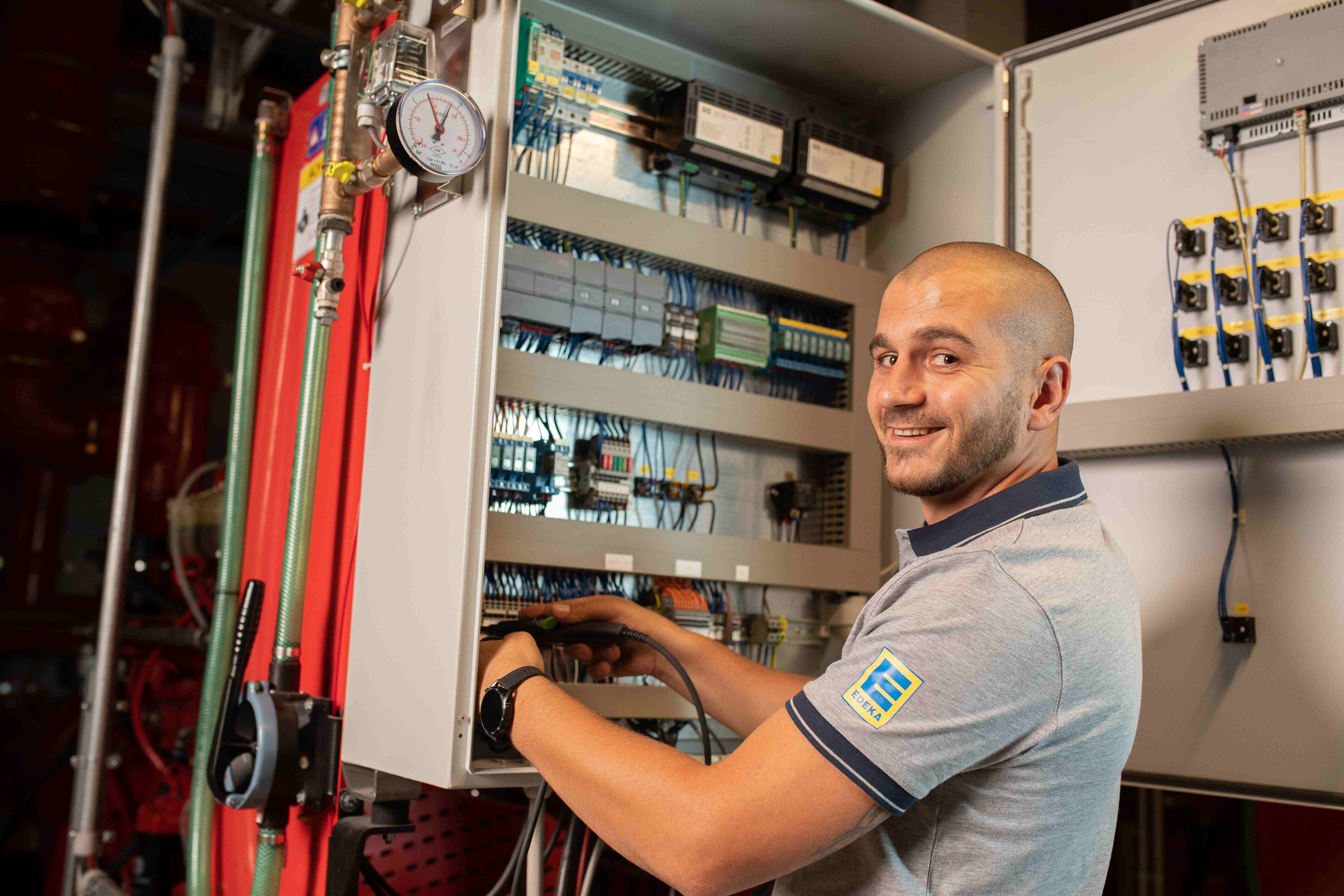 Mann mit sehr kurzen Haaren steht an einem geöffneten Verteilerschrank. Er trägt ein graues EDEKA Polo-Shirt und lächelt.
