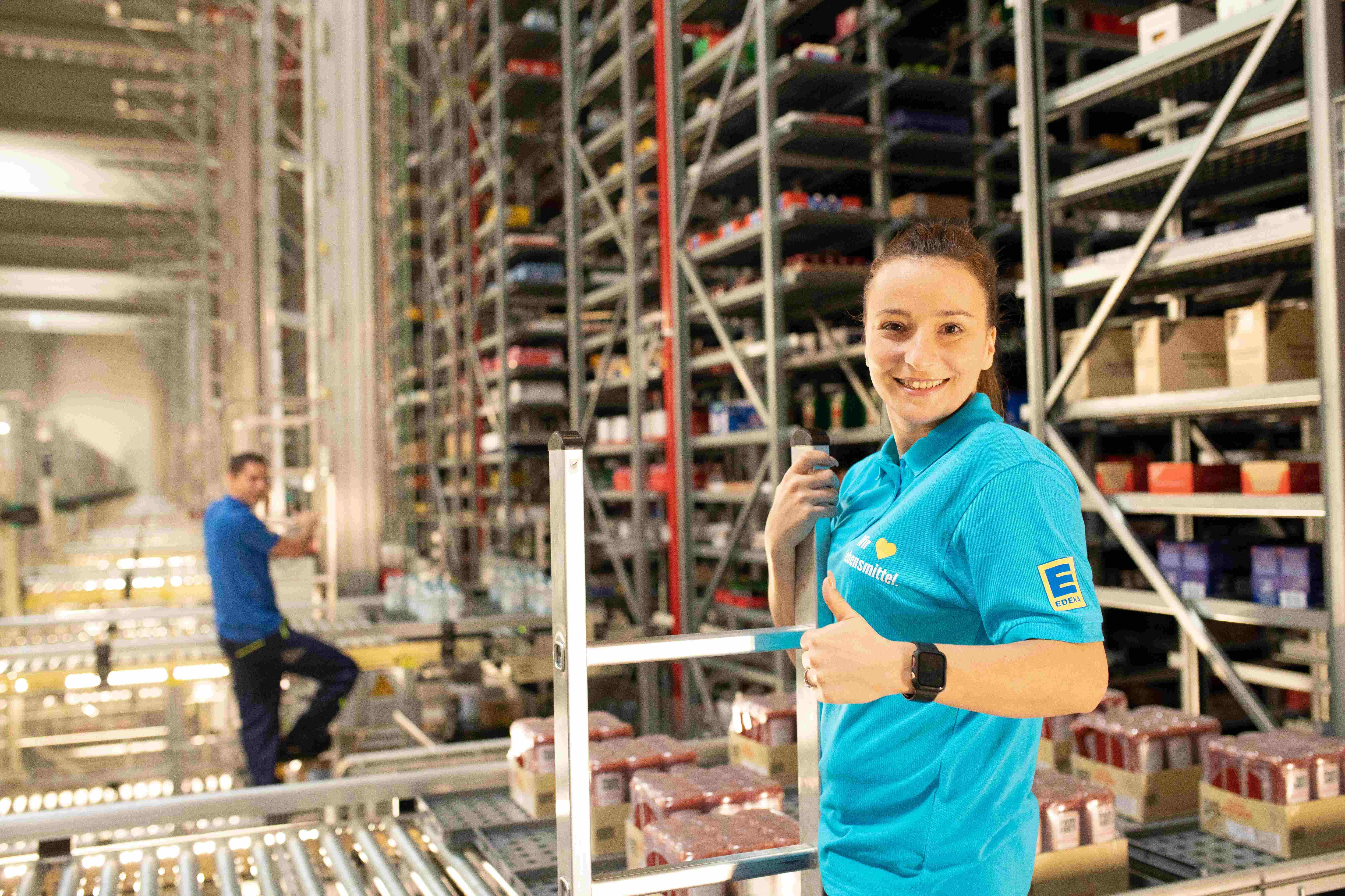 Eine Frau mit dunklen Haaren steht im Warenlager an einer Leiter. Sie trägt ein hellblaues Polo-Shirt mit EDEKA-Logo.