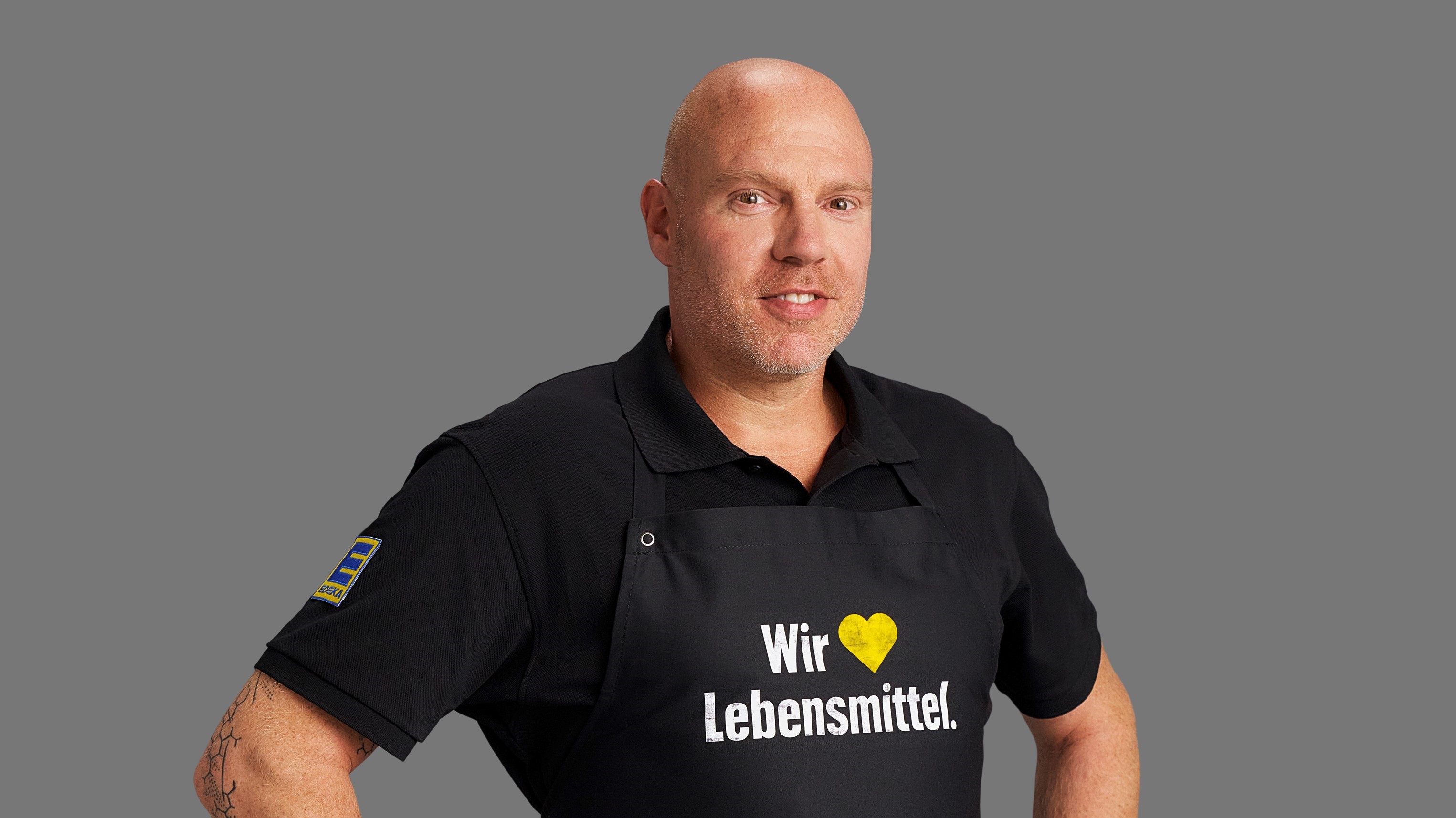 Robert Krieger vor grauem Hintergrund. Er trägt ein schwarzes Polo-Hemd und eine schwarze Schürze mit Aufschrift.