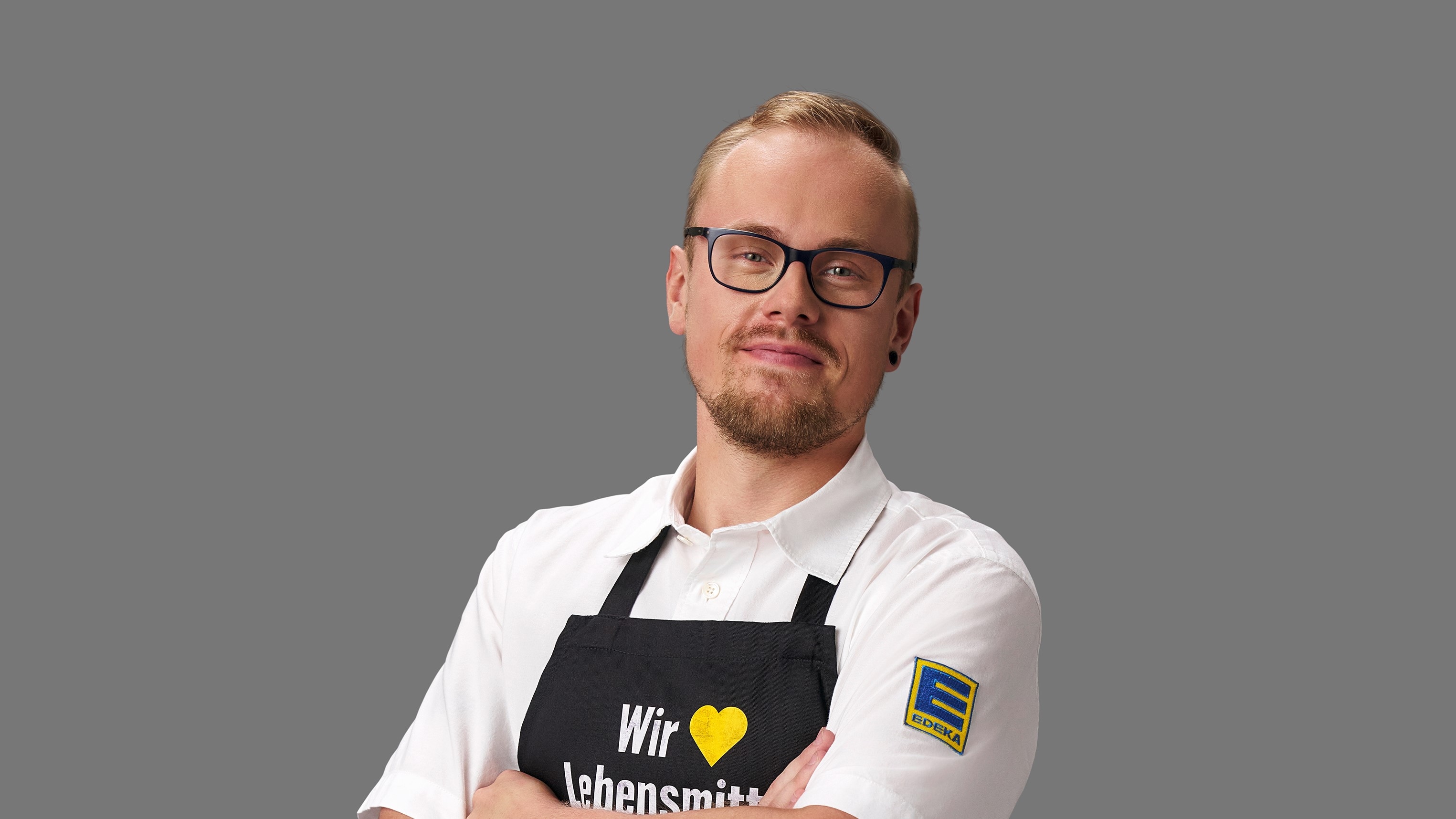 Rene Mattfeld vor einem grauen Hintergrund. Er trägt eine schwarze Brille, ein weißes EDEKA-Hemd und eine schwarze Schürze.