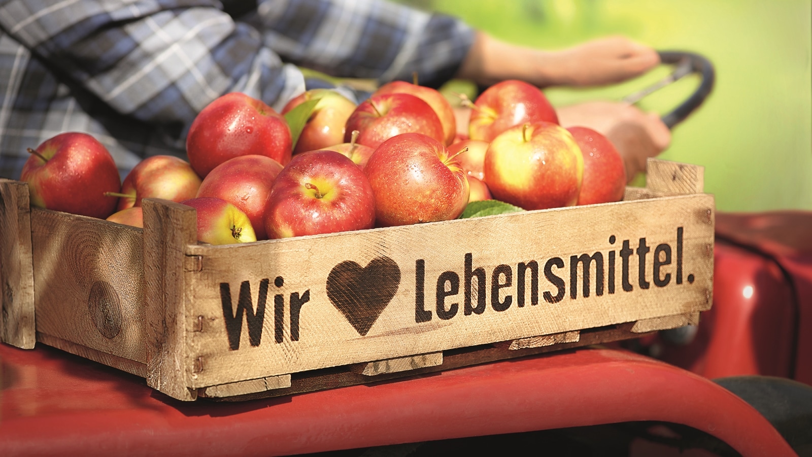 Ein Landwirt fährt einen roten Traktor, darauf steht eine Kiste mit Äpfeltn und der Aufschrift Wir lieben Lebensmittel.
