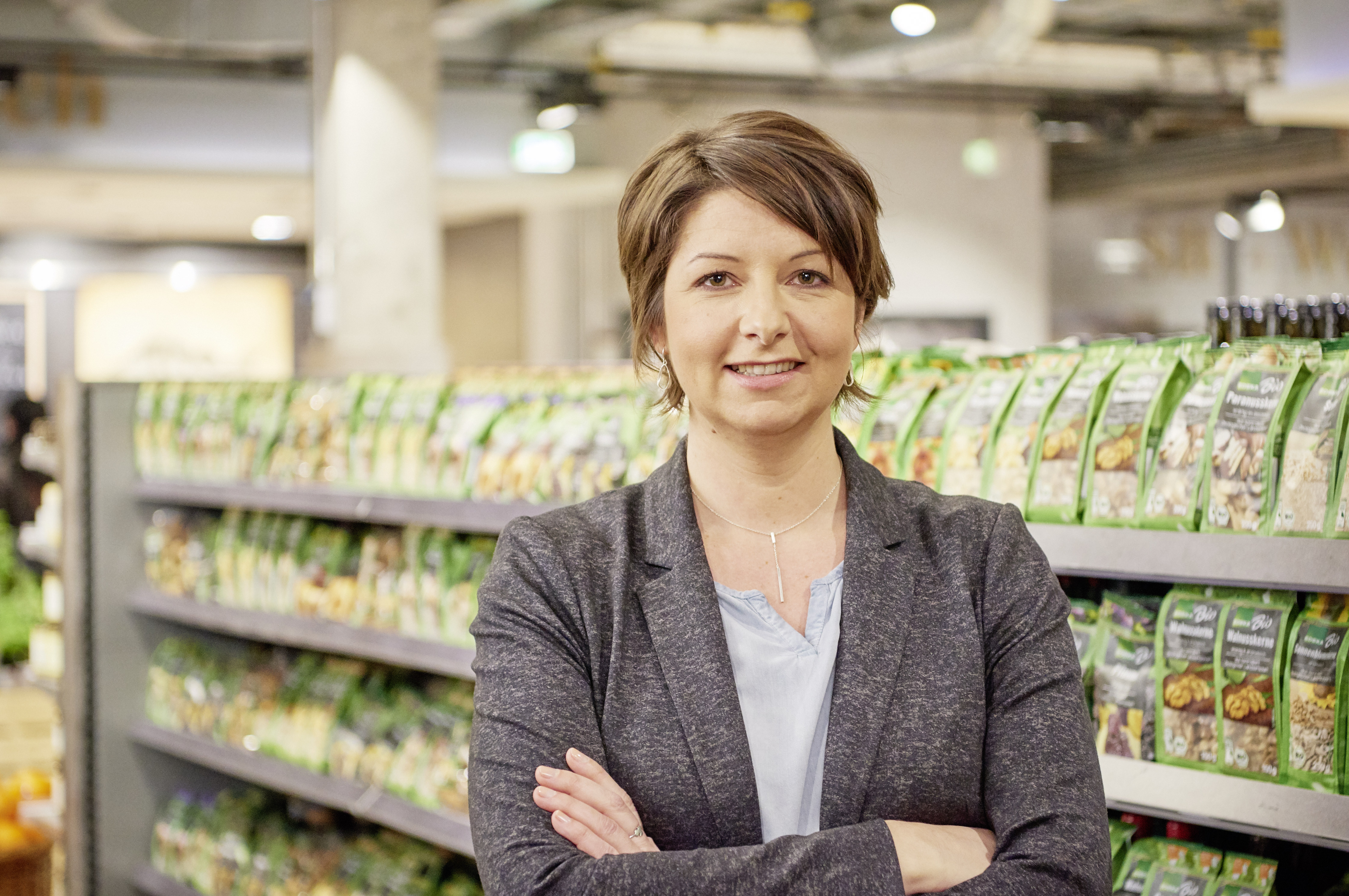 Frau im grauen Blazer steht mit verschränkten Armen vor dem Bio-Regal eines EDEKA-Marktes. 