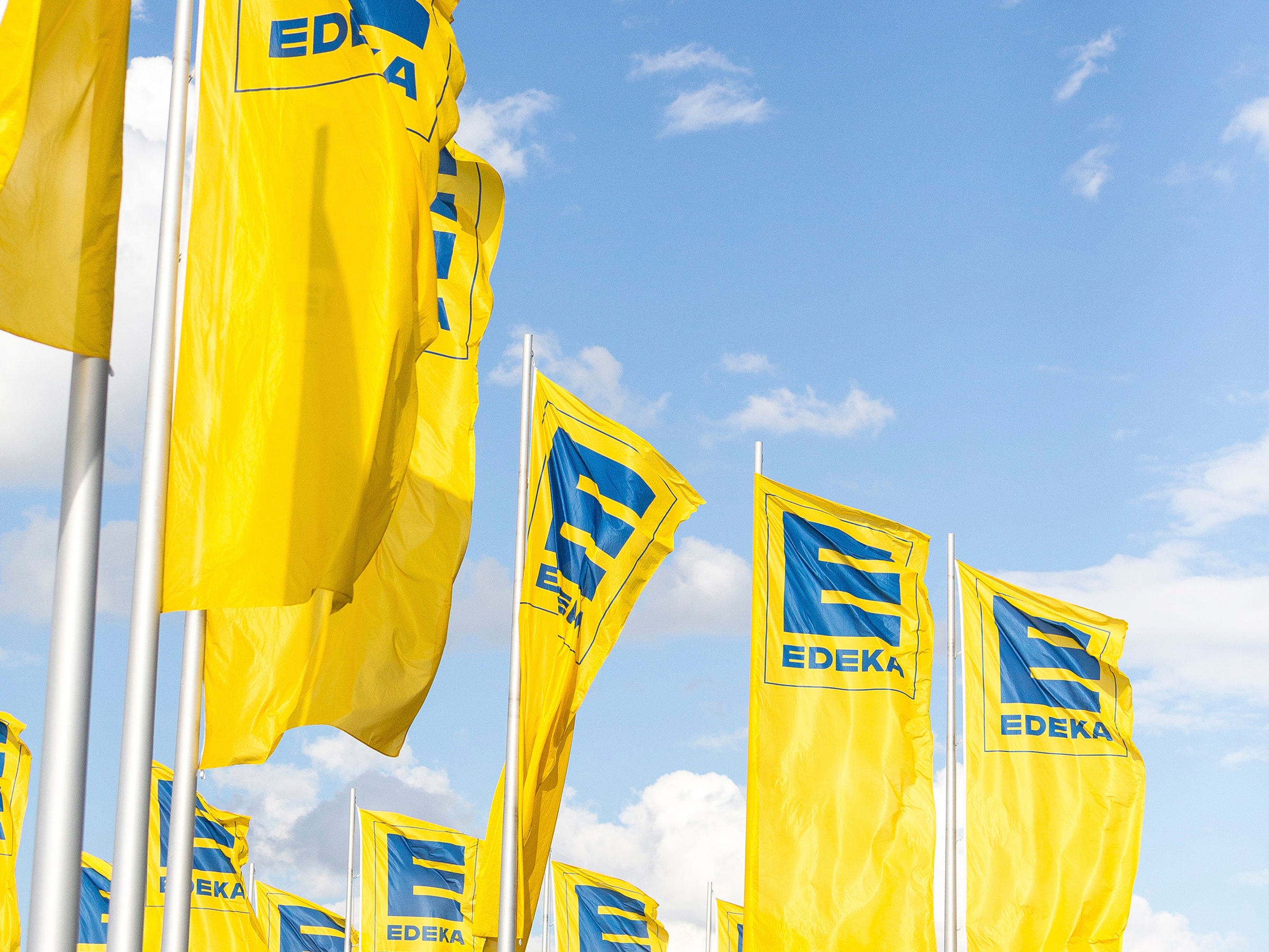 Gelbe Fahnen mit blauem EDEKA-Logo vor einem blauen Himmel mit weißen Wolken.