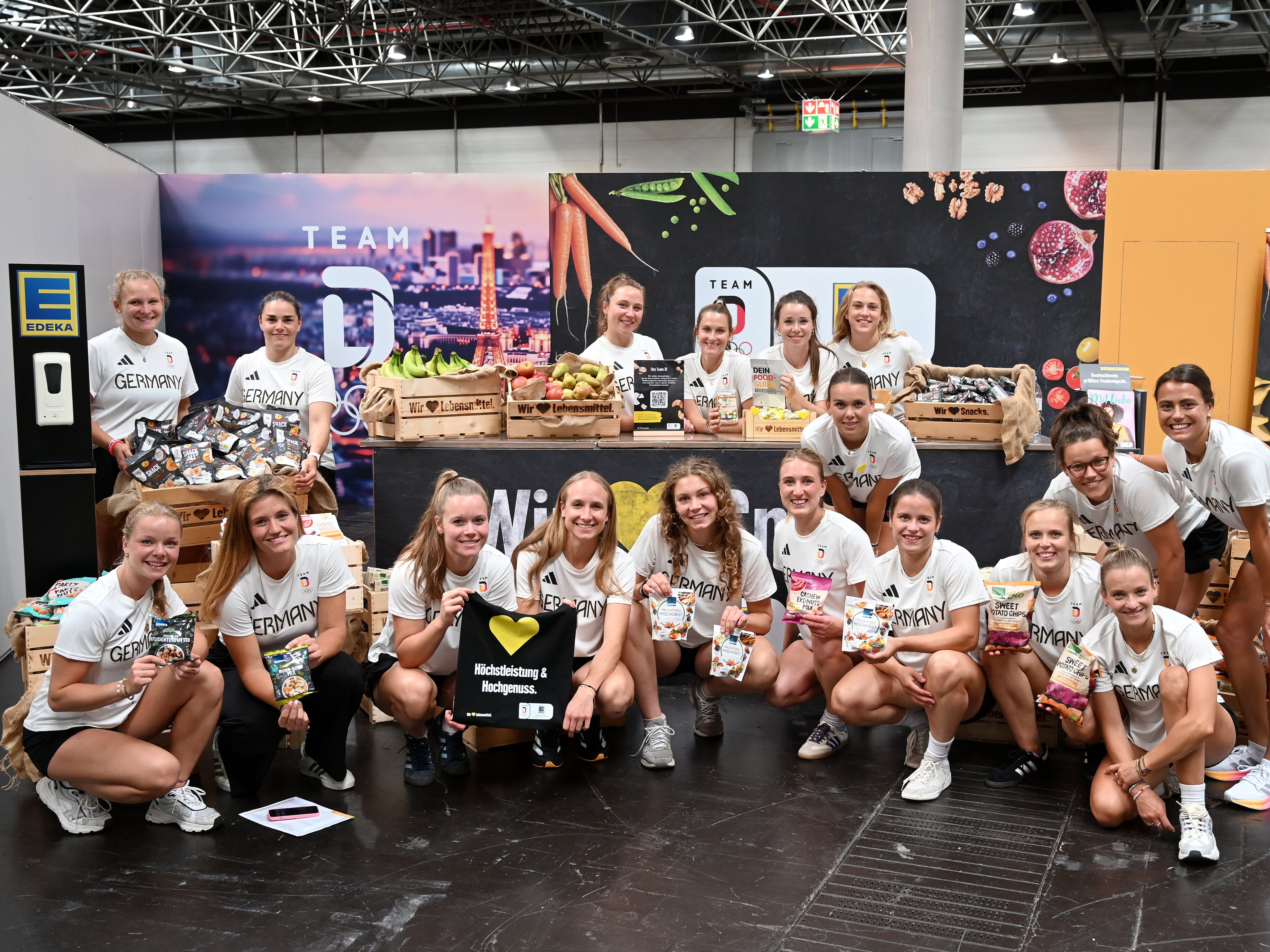 Eine Gruppe von rund 20 lächelnden Sportlerinnen posiert vor einem EDEKA-Messestand mit frischem Obst und Snacks. 