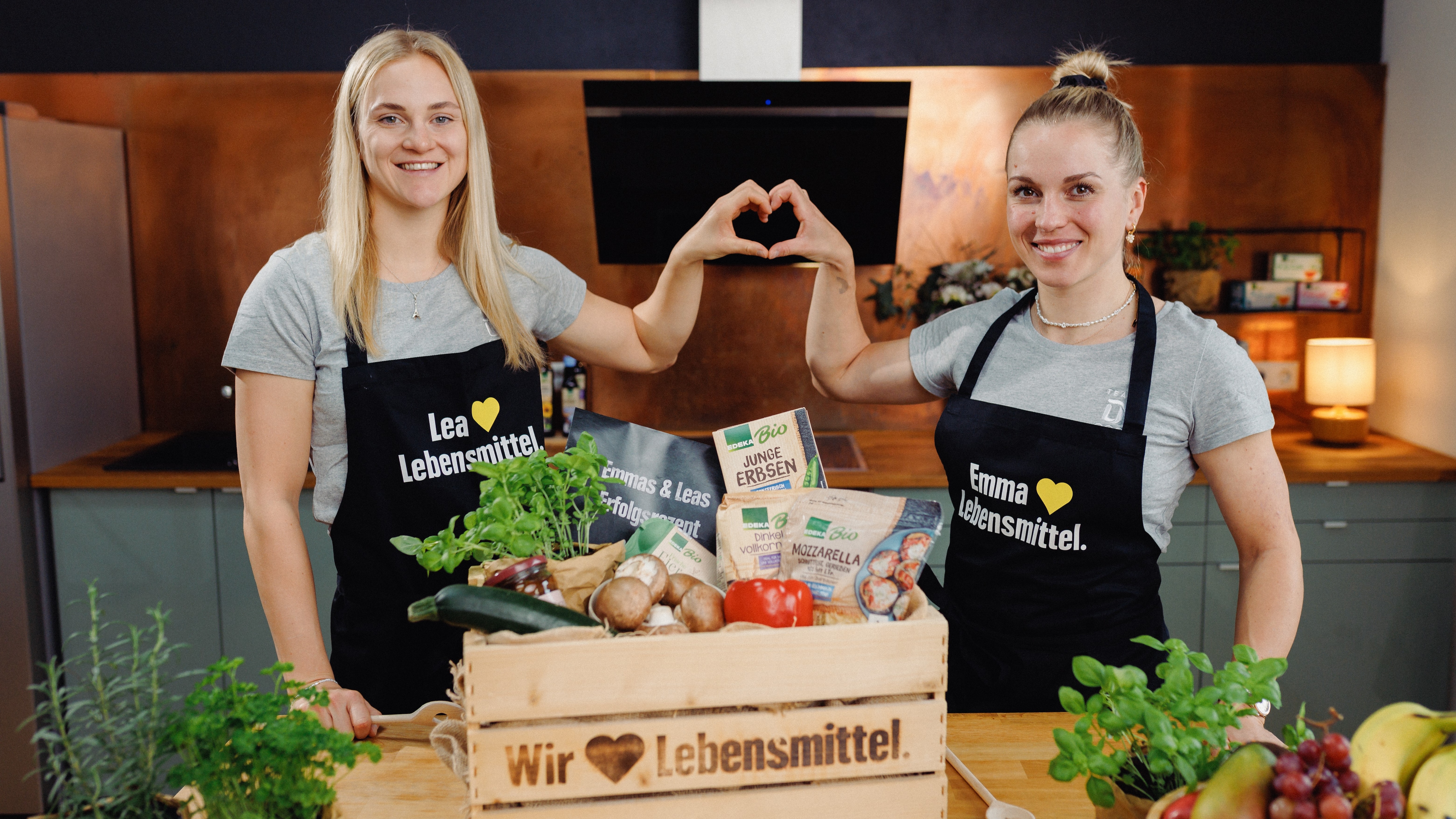 Zwei Sportlerinnen stehen in einer Küche und formen mit den Händen ein Herz. Vor ihnen steht eine Holzkiste mit Lebensmitteln