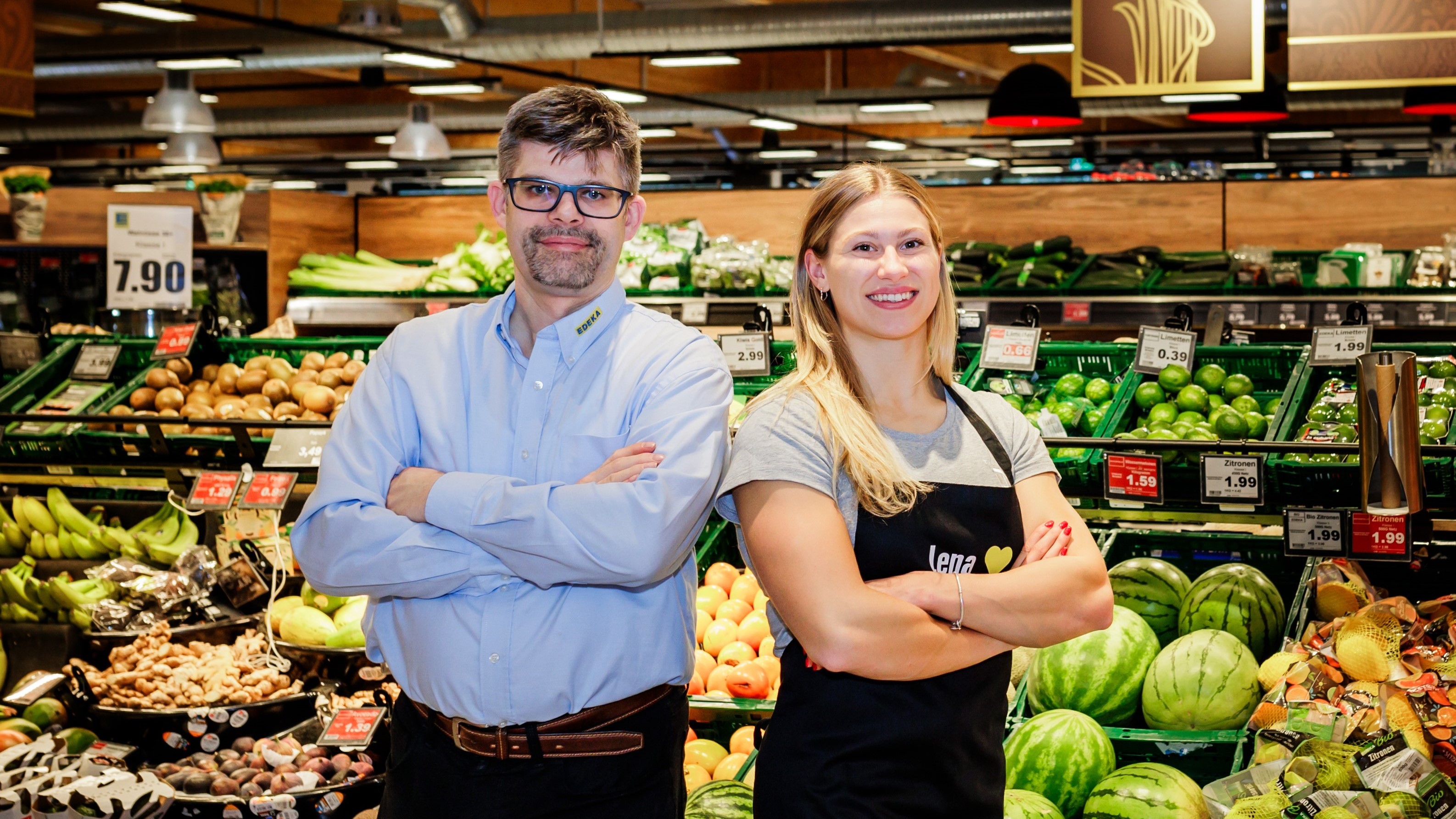 Ein Kaufmann und eine Athletin stehen lächelnd mit verschränkten Armen vor einem Gemüse-Regal in einem EDEKA-Markt.