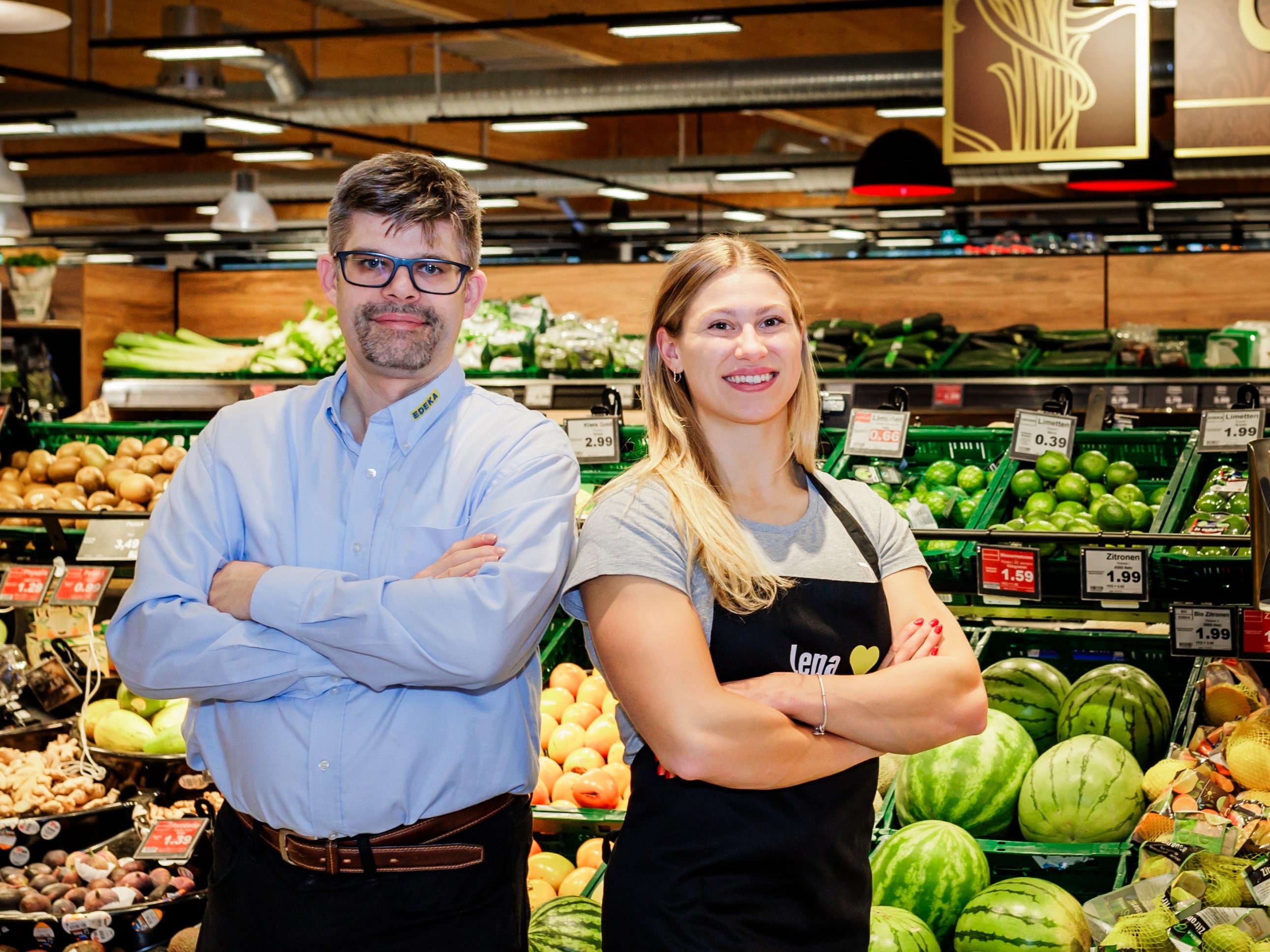 Ein Kaufmann und eine Athletin stehen lächelnd mit verschränkten Armen vor einem Gemüse-Regal in einem EDEKA-Markt.