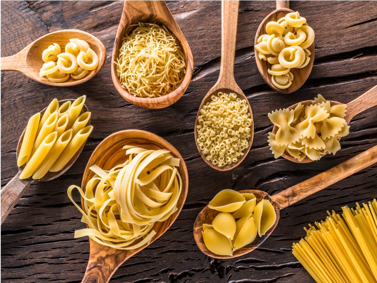 Auf einen dunklen Holztisch liegen Holzkochlöffel, gefüllt mit verschiedenen Pasta-Sorten.