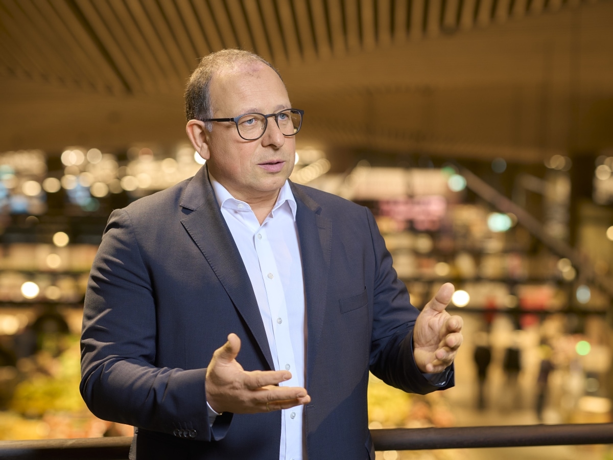 Ein Mann mit Brille, dunklem Jackett und weißem Hemd im Vordergrund, dahinter unscharf ein Lebensmittelmarkt