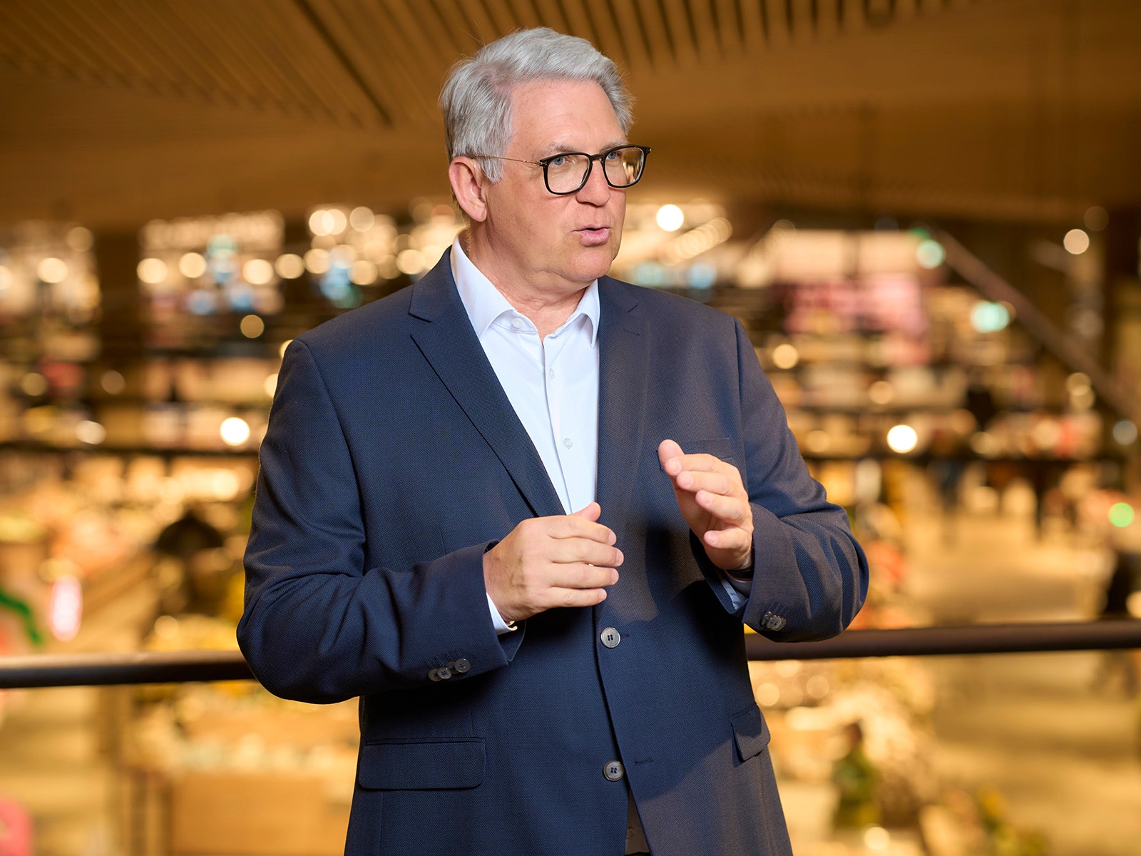 Portrait Uwe Kohler. Mann mit grauem Haar, trägt Brille weißes Hemd und Jackett. Er spricht uns gestikuliert mit den Händen. 