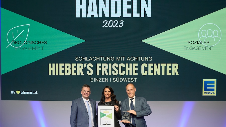 Auf einer Bühne stehen zwei Männer in Anzügen mit einer Frau in ihrer Mitte. Sie hält eine Urkunde.