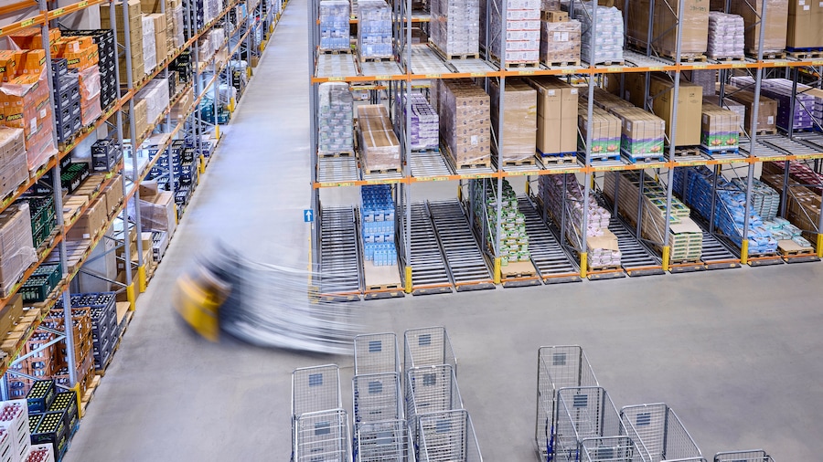 Großes Lager mit hohen Regalen voller verpackter Waren; leere Metallwagen im Vordergrund und ein Flurförderfahrzeug.