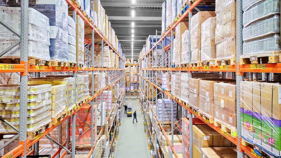 Blick in ein gut beleuchtetes Lager mit hohen Regalen voller Waren; zwei Personen im Gang, eine davon bedient einen Hubwagen.