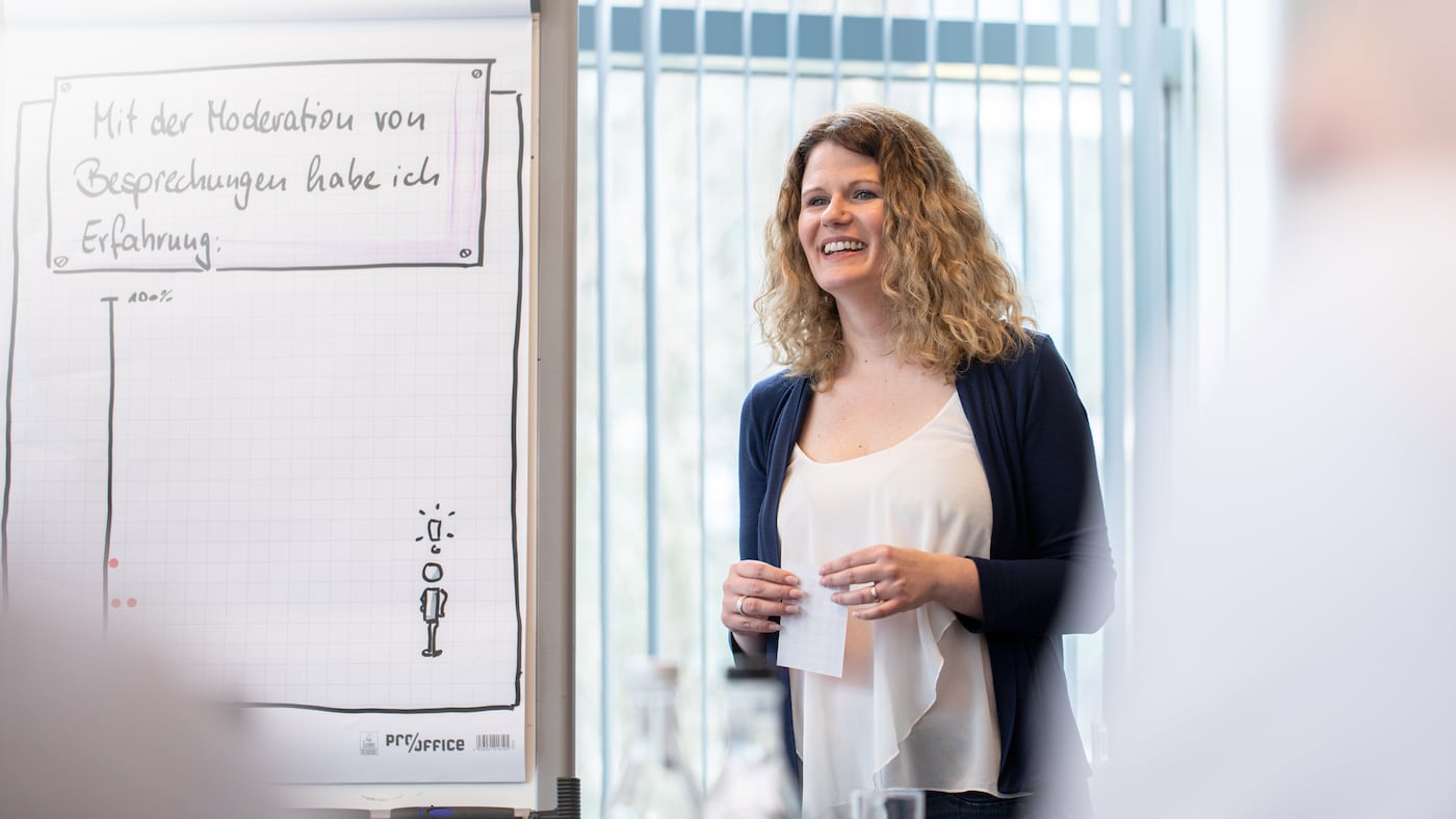 Junge Frau steht neben einem Flipchart und präsentiert während eines Seminars.