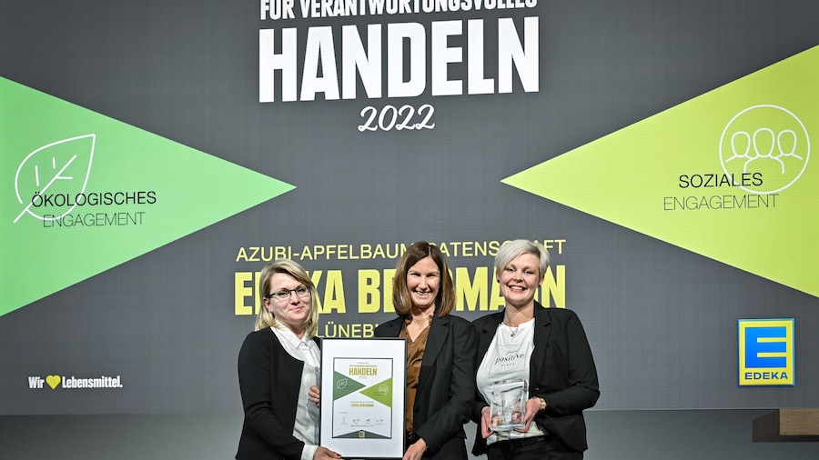 Drei Frauen stehen in festlicher Kleidung auf einer Bühne. Zwei halten eine gerahmte Urkunde, die dritte eine Glas-Trophäe. 