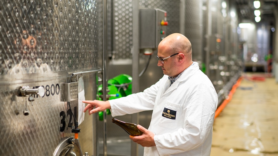 Mitarbeiter in einem weißen Kittel stellt die Abfüllanlage ein und hält eine Flasche Wein in der Hand.