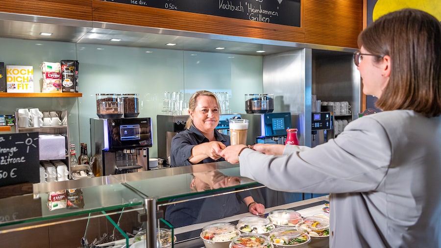 Ein blonde Frau reicht einen Latte Macchiato im Glas über den Tresen einer Kantine. 