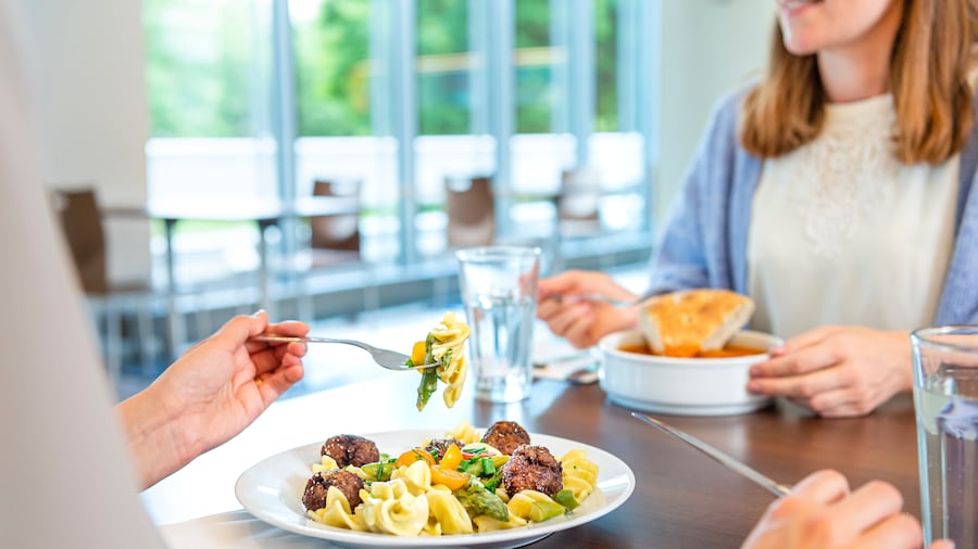 Zwei Personen sitzen gemeinsam an einem Tisch und essen. Im Vordergrund sieht man einen Teller mit Pasta, Gemüse und Falafel.