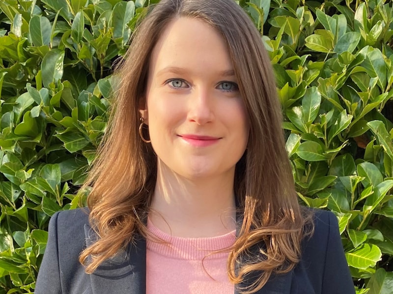 Eine junge Frau steht vor einer grünen Hecke. Sie hat lange, braune Haare und trägt ein dunkelblaues Sakko.