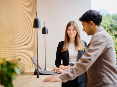 Eine junge Frau und ein junger Mann unterhalten sich an einem Stehtisch vor einer Fensterfront.