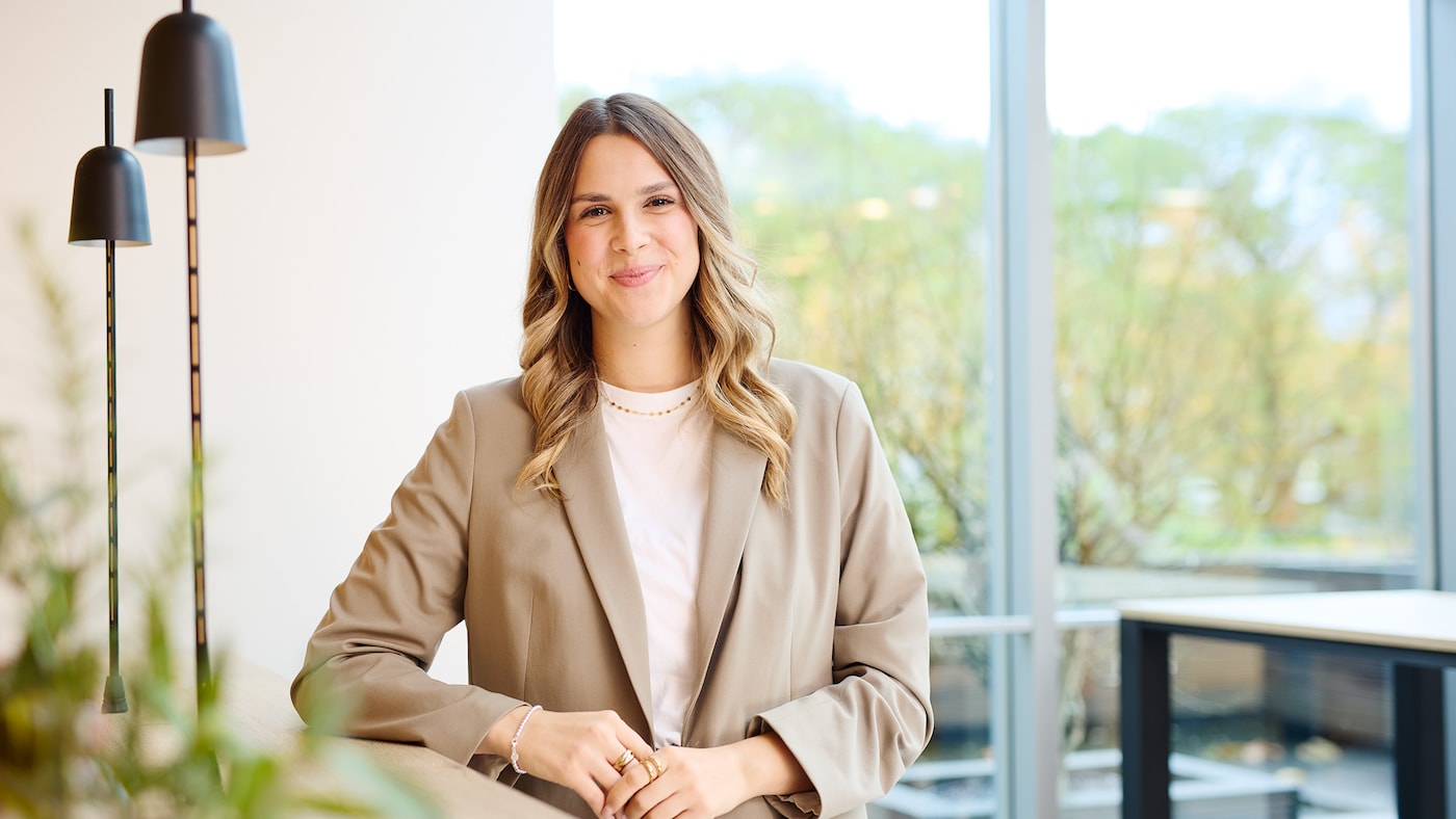 Frau in einem beigen Blazter lehnt an einen Holztisch mit zwei schwarzen Lampen und lächelt in die Kamera.