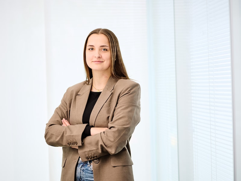 Katharina-Sophia Schulz steht mit verschränkten Armen vor einem hellen Hintergrund und lächelt in die Kamera.