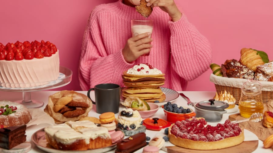Frau in pinkfarbenem Pulli sitzt vor einm reich gedeckten Tisch mit verschiedenem süßen Gebäck