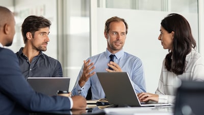 Vier Personen sitzen gemeinsam an einem Tisch und Arbeiten an Laptops, während sie sich austauschen.