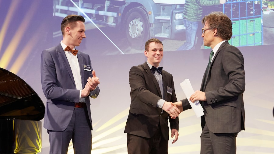 Drei Männer in Anzügen auf einer Bühne. Der linke klatscht, der mittlere und der rechts stehende schütteln Hände.