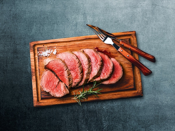 Ein Holzbrett, angerichtet mit gegarten Roastbeefscheiben, daneben liegen Messer und Gabel.