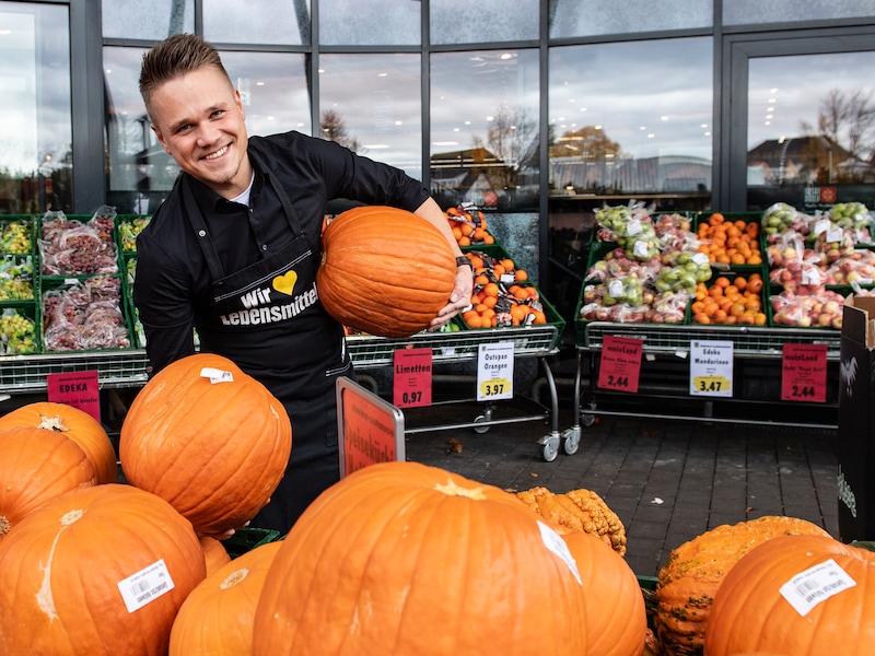 Marvin Waschkuhn vor einem EDEKA-Markt. Er hält in jeder Hand einen Riesenkürbis, trägt eine schwarze EDEKA-Schürze und lacht