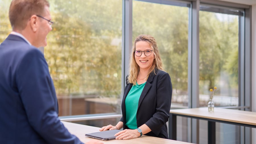 Zwei Personen stehen in einem modernen Büro an einem hellen Tisch. Die Frau trägt eine Brille und eine schwarze Jacke.
