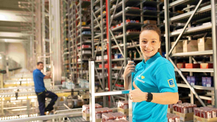 Eine Frau mit dunklen Haaren steht im Warenlager an einer Leiter. Sie trägt ein hellblaues Polo-Shirt mit EDEKA-Logo.