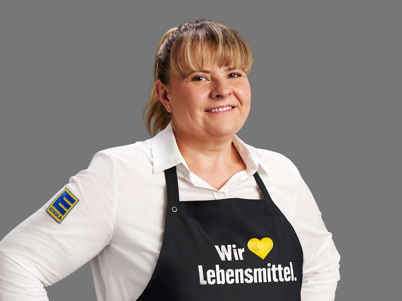 Karen Bich vor grauem Hintergrund. Die blonde Frau trägt ein weißes Hemd und eine schwarze Schürze mit Aufschrift.