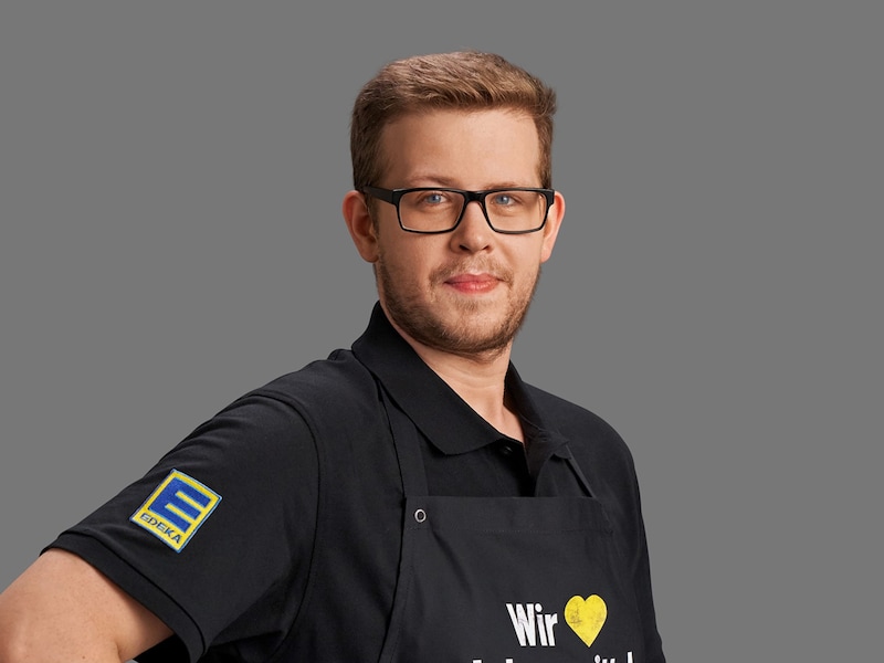 André Gruber vor grauem Hintergrund. Er trägt eine eckige Brille, ein schwarzes Polo-Shirt und eine schwarze Schürze.