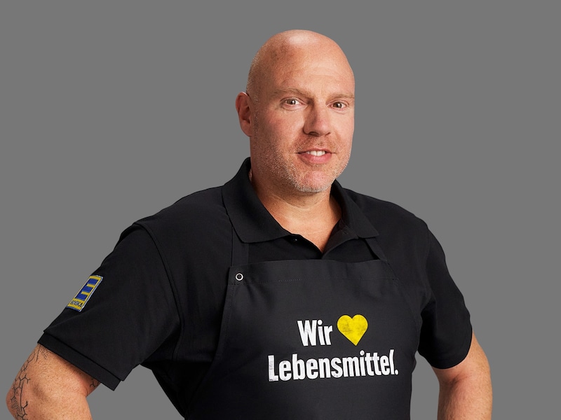 Robert Krieger vor grauem Hintergrund. Er trägt ein schwarzes Polo-Hemd und eine schwarze Schürze mit Aufschrift.