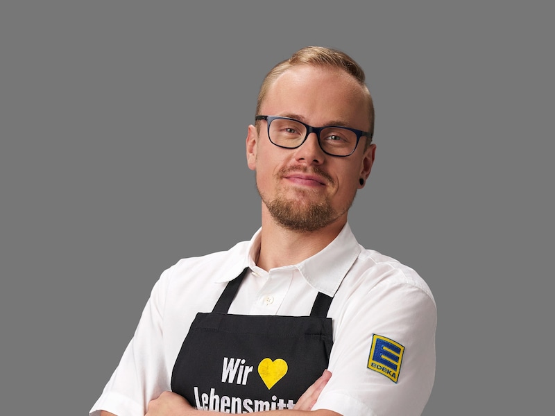 Rene Mattfeld vor einem grauen Hintergrund. Er trägt eine schwarze Brille, ein weißes EDEKA-Hemd und eine schwarze Schürze.
