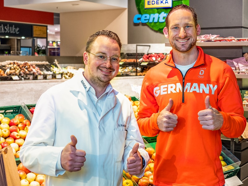 Ein Athlet und ein Kaufmann stehen vor einem Gemüseregal im EDEKA-Markt. Beide lächeln und zeigen die Daumen nach oben.