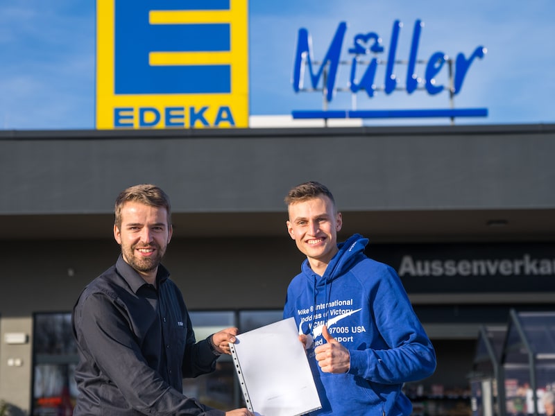 Ein Athlet und ein EDEKA-Kaufmann stehen gemeinsam vor einem EDEKA-Markt und lächeln in die Kamera.