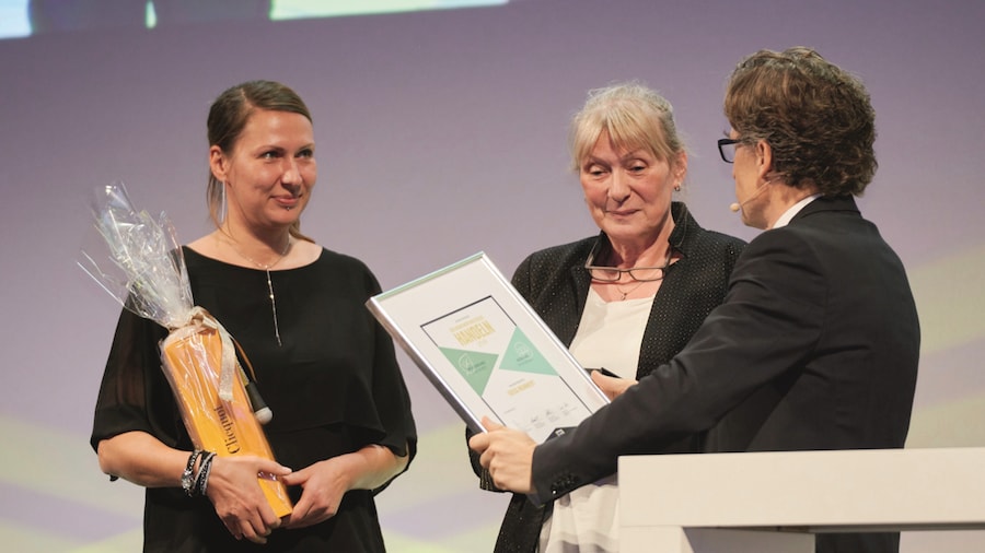 Eine ältere und eine jüngere Frau bekommen eine gerahmte Urkunde von einem Herrn im dunklen Anzug überreicht. 