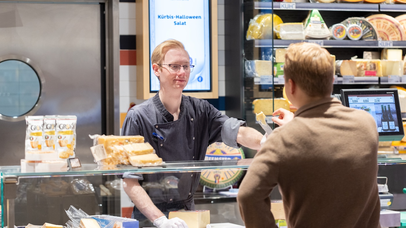 Mitarbeiter an der Bedientheke reicht Käse an