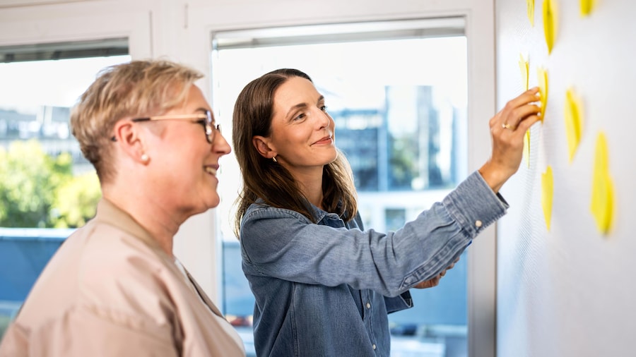 Kolleginnen kleben Postits an eine Wand und sprechen darüber