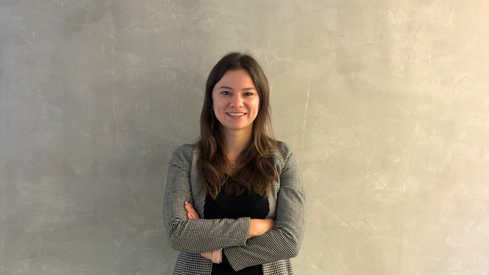 Katarina Hamann steht vor einer Wand in Betonoptik und lächelt mit verschränkten Abend in die Kamera. 